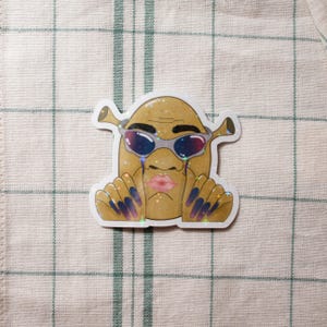 May include: A sticker featuring a cartoon character with a potato-like body, Shrek-like ears, and sunglasses. The character has a pink pouty lip, blue and purple nails, and tears. The sticker has a glittery, holographic effect.