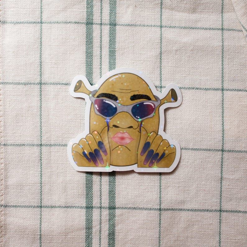 May include: A sticker featuring a cartoon character with a potato-like body, Shrek-like ears, and sunglasses. The character has a pink pouty lip, blue and purple nails, and tears. The sticker has a glittery, holographic effect.