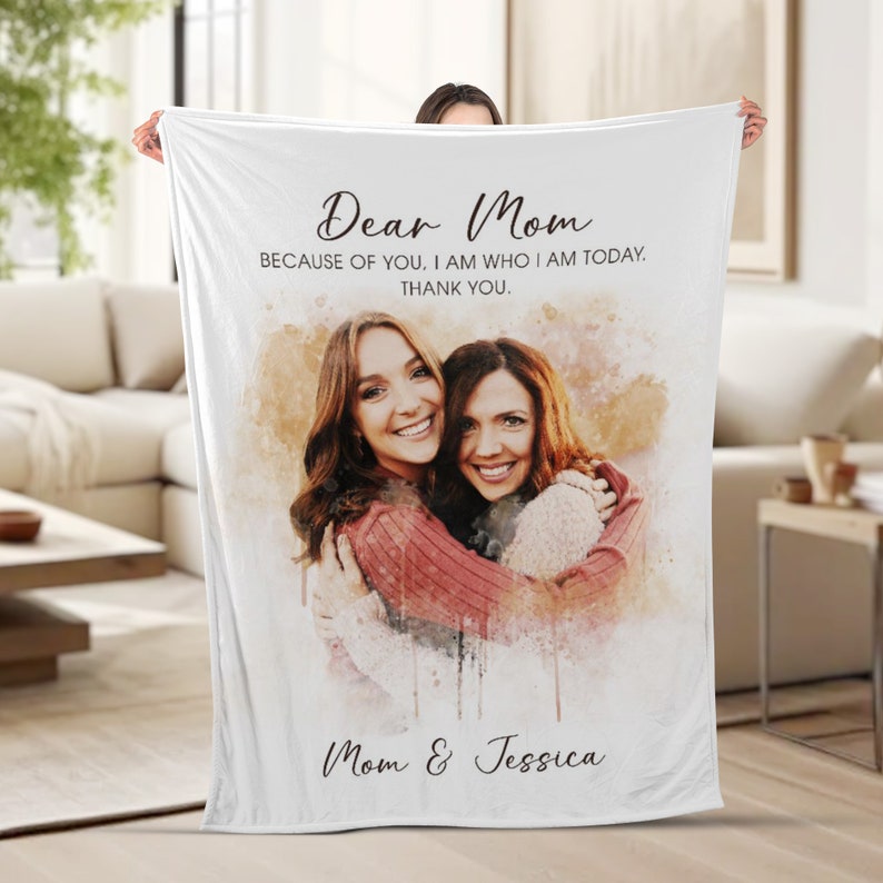 K&ouml;nnte beinhalten: Eine wei&szlig;e Fleece-Decke mit einem Aquarellbild von zwei Frauen, die sich umarmen. Der Text "Dear Mom" befindet sich oben auf der Decke, gefolgt von "Because of you, I am who I am today. Thank you." Der Text "Mom & Jessica" befindet sich unten auf der Decke.