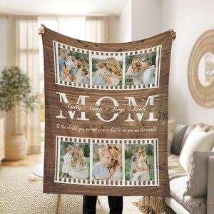Op de afbeelding: Een bruine houten deken met een witte rand en een wit 'MOM'-ontwerp. De deken heeft een collage van foto's in een filmstripontwerp. De tekst 'To the world you are one person, but to us you are the world' staat onder de foto's gedrukt. De namen 'Emily', 'Emma', 'Jorge' en 'Noah' staan in cursief boven het 'MOM'-ontwerp.