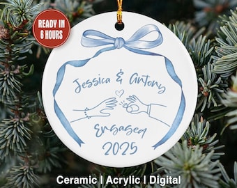 May include: A white ceramic ornament with a blue ribbon design and the text "Jessica & Antony Engaged 2025." The ornament has a gold string for hanging and is surrounded by evergreen branches. The text "Ready in 6 hours" is also visible.