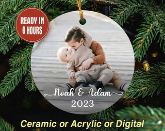 May include: A round Christmas ornament featuring a photo of two children. The ornament has the names "Noah & Adam" and the year "2023" printed on it. The text "Ready in 6 hours" is in a red circle. The bottom text says "Ceramic or Acrylic or Digital".
