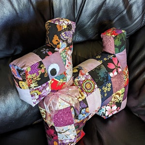 May include: A patchwork dog cushion with a pink ribbon. The cushion is made of various fabrics with floral and geometric patterns in shades of purple, pink, black and gold.