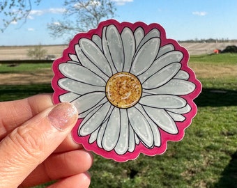 May include: A white daisy with a yellow center, surrounded by a pink border. The daisy is drawn in a cartoon style and has a glitter-like texture.