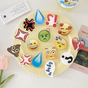 May include: A white plate with a variety of colorful emoji-themed stickers. The stickers feature different expressions, including happy, sad, angry, sleepy, and surprised. There is also a sticker of a poop emoji, a question mark, and a heart.