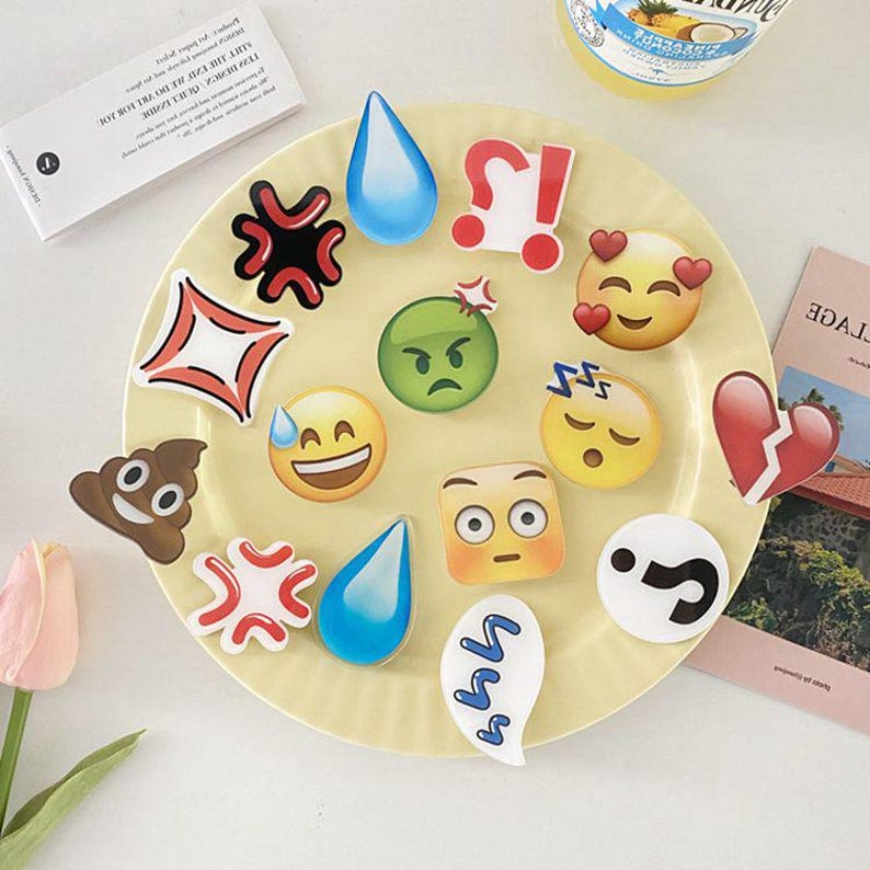 May include: A white plate with a variety of colorful emoji-themed stickers. The stickers feature different expressions, including happy, sad, angry, sleepy, and surprised. There is also a sticker of a poop emoji, a question mark, and a heart.
