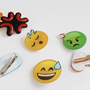 May include: Six colorful plastic hair clips with different emoji faces. The clips are shaped like a heart, a teardrop, and circles. The emojis include a sleeping face, a frowning face, a smiling face with a sweat drop, and an angry face.