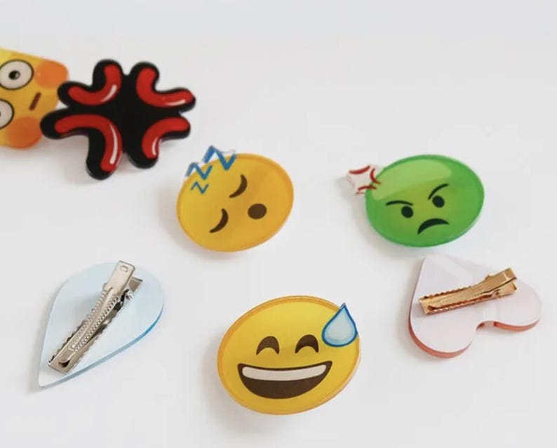 May include: Six colorful plastic hair clips with different emoji faces. The clips are shaped like a heart, a teardrop, and circles. The emojis include a sleeping face, a frowning face, a smiling face with a sweat drop, and an angry face.