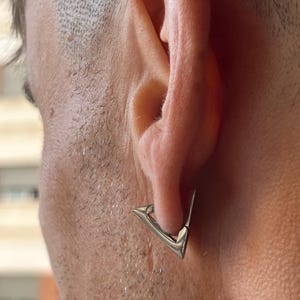 Può includere: Un orecchino a forma di triangolo argentato con una punta appuntita. L'orecchino è indossato sul lobo dell'orecchio sinistro.