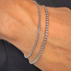 Peut inclure: Deux bracelets en chaîne argentée sur le poignet d'une personne. Les bracelets sont faits de petites perles rondes.