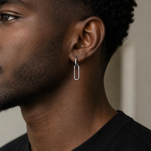 Puede incluir: Un pendiente de aro rectangular plateado se lleva en la oreja. El pendiente tiene un diseño sencillo y moderno con un acabado pulido. La persona lleva una camiseta negra.