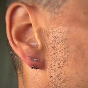 Puede incluir: Primer plano de la oreja de una persona con tres pequeños pendientes de plata. Los pendientes tienen forma de línea curva, círculo pequeño y círculo pequeño con un pequeño diamante.