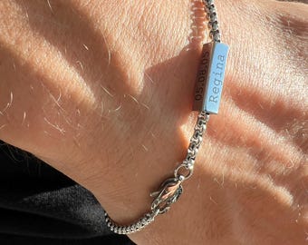 Puede incluir: Una pulsera plateada con un dije rectangular azul grabado con el nombre "Regina" y la fecha "05.08.05". La pulsera presenta un diseño de cadena de caja y un cierre decorativo.