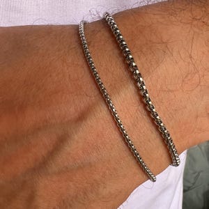 May include: Two silver chain bracelets on a person's wrist. The bracelets are made of small, round links.