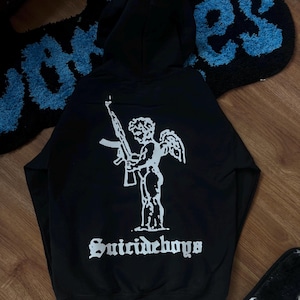 Può includere: Felpa nera con grafica bianca di un angelo che tiene un fucile e la scritta "Suicideboys" in carattere gotico. La felpa ha un cappuccio. Un tappeto blu e nero è sullo sfondo.