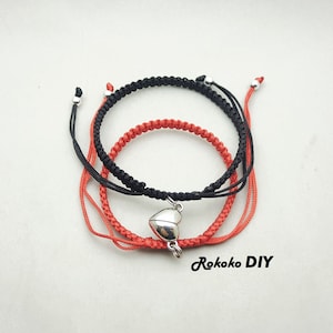 Op de afbeelding: Twee vriendschapsarmbanden, een zwarte en een rode, met een zilveren hartbedel. De armbanden zijn met een knoop aan elkaar gebonden.