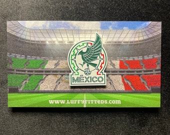 May include: A metal pin featuring a green eagle emblem above a soccer ball and the text MÉXICO, displayed on a rectangular card showing a stadium with green, white, and red seating sections and the website text WWW.LUFFYFITTEDS.COM at the bottom.