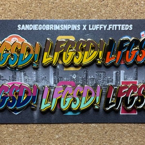 May include: Six gold-colored pins with the text "LIFGSD!" in a rainbow color scheme. The pins are arranged in two rows of three. The text "SANDIEGOBRIMSPIN X LUFFY.FITTEDS" is printed on a white background behind the pins.
