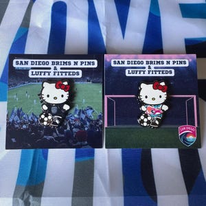 May include: Two enamel pins featuring Hello Kitty in soccer uniforms, holding a soccer ball. The pins are on cards with the text "SAN DIEGO BRIMS N PINS & LUFFY FITTEDS". One pin has a black uniform, the other a teal and pink uniform. The San Diego logo is on one card.