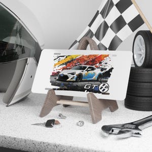 May include: White license plate with a colorful graphic of a white and blue sports car, the text "GT" and a checkered flag. The plate is displayed on a wooden stand, with a helmet, tires, and tools in the background.