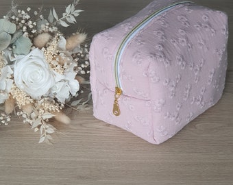 May include: A light pink, square cosmetic bag with a floral pattern. The bag has a gold zipper and pull, and a white interior. A bouquet of white flowers is in the background.