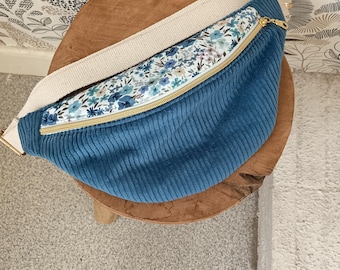 Peut inclure: Sac banane en velours côtelé bleu sarcelle avec un imprimé floral et une sangle crème. Le sac est doté d'une fermeture éclair dorée et est présenté sur une surface en bois. Dimensions: environ 30 cm de long.