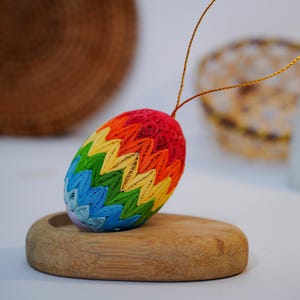 Puede incluir: Adorno de huevo de Pascua colorido y hecho a mano. El huevo está hecho con quilling de papel en colores del arcoíris, incluyendo rojo, naranja, amarillo, verde, azul y morado. Tiene una cuerda dorada para colgar y está sobre un pequeño soporte de madera.