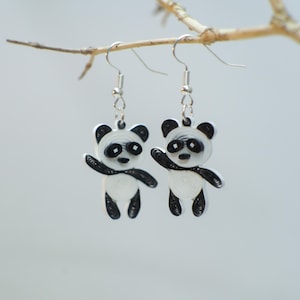 May include: A pair of dangling earrings featuring panda bear charms crafted from coiled paper strips. The pandas are white with black ears, eye patches, arms, and legs. Each charm hangs from a silver-toned metal fishhook ear wire.