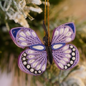 May include: A detailed butterfly ornament made using paper quilling techniques. The butterfly is predominantly purple, with white and black accents, and a gold hanging string. The wings display a layered design, giving a textured look. This decorative piece is ideal for adding a natural touch to any room.