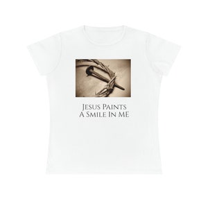 Puede incluir: Camiseta blanca con una imagen en sepia de un clavo en una corona de espinas. El texto "JESUS PAINTS A SMILE IN ME" está impreso debajo de la imagen.