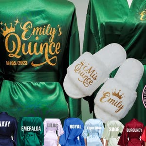 May include: Two green satin robes with gold lettering. One robe says "Emily's Quince 08/05/2023". The other robe says "Emily". Two white slippers with gold lettering. One slipper says "Mis Quince". The other slipper says "Emily".
