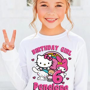 May include: A white long-sleeve shirt featuring a colorful graphic of Hello Kitty, My Melody, and other characters, with the text "BIRTHDAY GIRL", the number "6", and the name "Penelope" in pink.