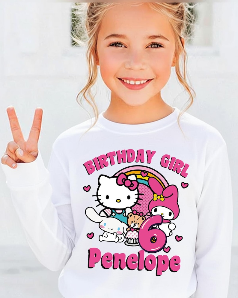May include: A white long-sleeve shirt featuring a colorful graphic of Hello Kitty, My Melody, and other characters, with the text "BIRTHDAY GIRL", the number "6", and the name "Penelope" in pink.