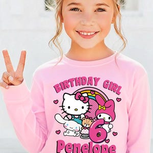 May include: A pink long-sleeved sweatshirt featuring a Hello Kitty design with a rainbow, the text "BIRTHDAY GIRL", the number "6", and the name "Penelope". The sweatshirt is worn by a young girl.