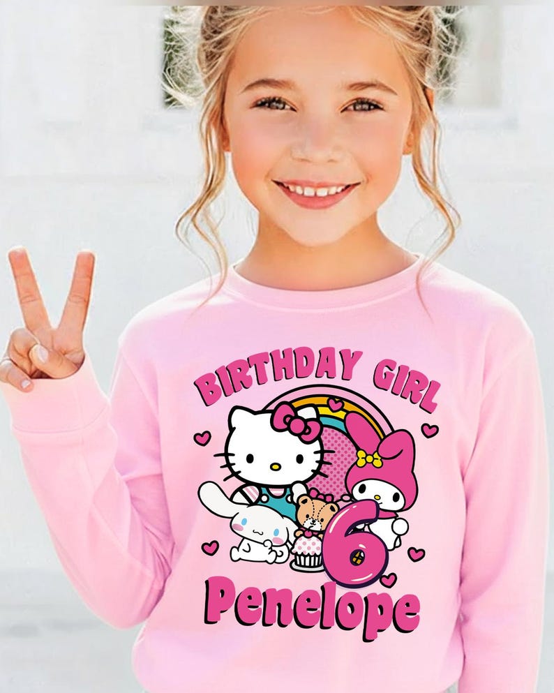 May include: A pink long-sleeved sweatshirt featuring a Hello Kitty design with a rainbow, the text "BIRTHDAY GIRL", the number "6", and the name "Penelope". The sweatshirt is worn by a young girl.