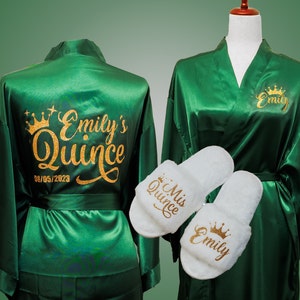 May include: Green satin robe with gold glitter lettering that says "Emily's Quince 08/05/2023" on the back. A matching robe with the name "Emily" and a crown is shown on a mannequin. Two white slippers with gold glitter lettering are included. One slipper says "Mis Quince" and the other says "Emily" with a crown.