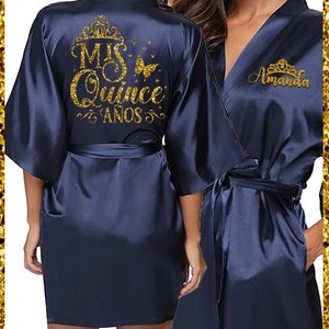 May include: Navy blue satin robe with gold glitter lettering on the back that says "Mis Quince Anos" with a crown and butterfly. The front of the robe has gold glitter lettering that says "Amanda" with a crown.