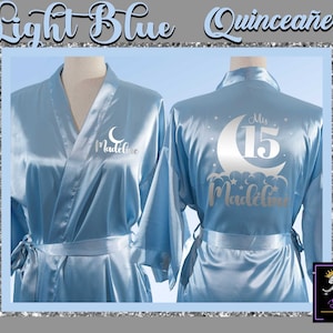 May include: Light blue satin robe with silver embroidery. The robe features a crescent moon with stars and the text "Mis 15 Madeline".