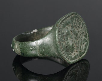 May include: A green bronze ring with a square-shaped bezel featuring an engraved design. The ring is worn and has a patina.