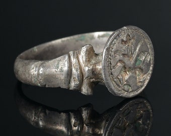 May include: A silver ring with a round, engraved bezel. The bezel features a stylized design of a bird with its wings spread.