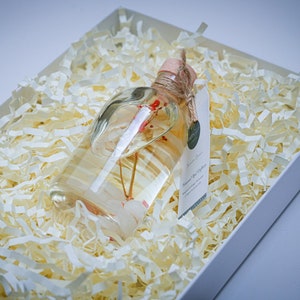 May include: A clear glass bottle with a cork stopper and a white label. The bottle is filled with a clear liquid and contains a small, dried flower. The bottle is sitting in a white box filled with white shredded paper.