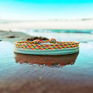 Könnte beinhalten: Ein farbenfrohes Armband mit einem Regenbogen-Seil-Design, das auf einer hellblauen Basis ruht. Das Armband befindet sich an einem Sandstrand mit dem Meer im Hintergrund. Das Seil besteht aus roten, gelben, grünen und blauen Fäden.