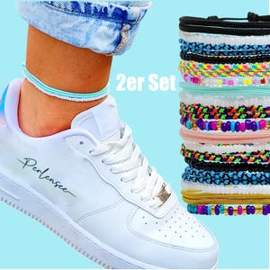 Puede incluir: Una pila de tobilleras coloridas con cuentas y trenzadas junto a una zapatilla blanca con la palabra "Perlensee" en el lateral. El texto "2er Set" es visible. Una tobillera se lleva en el tobillo. Las tobilleras son de varios colores.