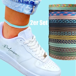 Puede incluir: Una zapatilla blanca con la palabra "Perlensee" en cursiva. Se lleva una tobillera azul claro con la zapatilla. Una pila de cordones trenzados de colores está a la derecha. El texto "2er Set" es visible.
