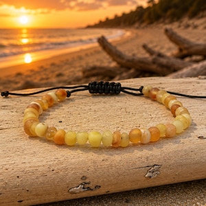 Könnte beinhalten: Ein Armband mit einer Reihe kleiner, abgerundeter, durchscheinender Perlen in verschiedenen Gelb-, Orange- und Blassgoldtönen. Die Perlen sind auf eine dünne schwarze Kordel gefädelt, die einen verstellbaren Schiebeknotenverschluss besitzt.