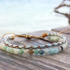 Könnte beinhalten: Zwei Armbänder mit Strandmotiv. Ein Armband hat hellblaues Leder mit braunen Nähten. Das andere Armband besteht aus hellgrünen und blauen Perlen. Die Armbänder liegen auf einer Holzoberfläche mit verschwommenem Ozeanhintergrund.