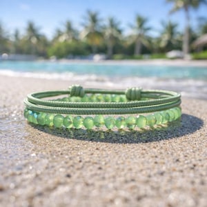 Könnte beinhalten: Zwei grüne Armbänder liegen auf einem Sandstrand. Das obere Armband besteht aus einem geflochtenen Kordel, das untere aus kleinen, facettierten grünen Perlen. Der Hintergrund zeigt eine tropische Strandszene mit Palmen und blauem Wasser.