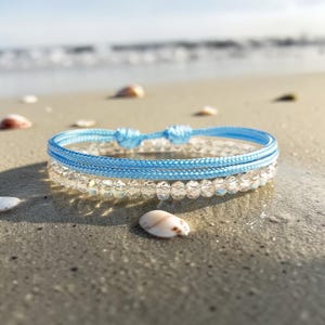 Könnte beinhalten: Zwei Armbänder an einem Sandstrand. Das obere Armband ist hellblau, aus geflochtenem Kordel und mit einem Knoten gebunden. Das untere Armband ist transparent, aus kleinen, runden Perlen. Muscheln sind um die Armbänder verstreut.