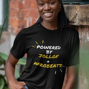 May include: A person wearing a black t-shirt with the text "Powered by Jollof + Afrobeats" in yellow. The person is standing in front of a brick wall.