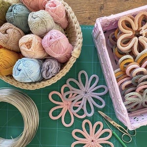Può includere: Una collezione di materiali per l'artigianato tra cui gomitoli di lana in vari colori pastello, sottobicchieri all'uncinetto a forma di fiore, un cestino, un contenitore di plastica rosa, forbici e una bobina di filo. Disposti su un tappetino verde.